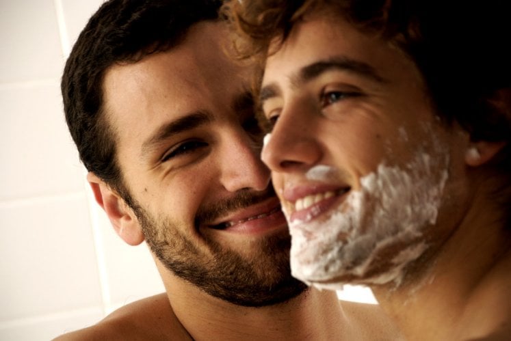 um jovem branco com espuma de barbear no rosto é observado por outro. Ambos sorriem.